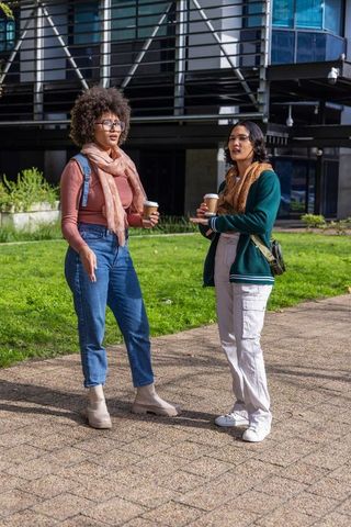 Campus friends chatting over coffee on university walkway with backpacks and scarves