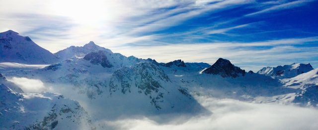 Snowcapped mountain range under clear sunny sky with clouds filling the valley below. Perfect for winter sport advertisements, nature wallpapers, travel brochures, or inspirational posters. Highlights the serene beauty of alpine scenery.