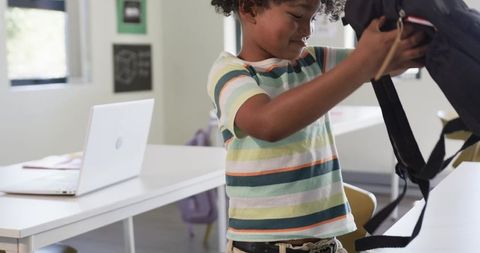 Young Student Packing Backpack in Bright Modern Classroom with Laptop Desk