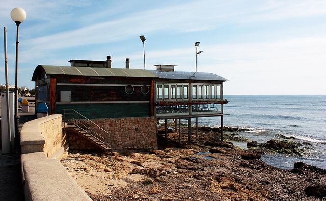 Rustic beachside restaurant with wooden and stone elements, overlooking the calm ocean. Ideal for dining concepts, vacation, relaxation, and coastal lifestyle promotions.