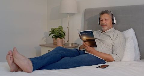 Senior Man Relaxing in Bed Listening to Music with Book