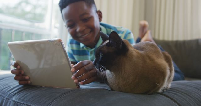 Happy african american boy lying on couch using tablet,stroking siamese pet cat and smiling. spending free time at home.