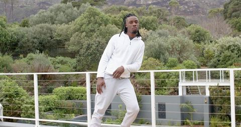 African American man leaning on balcony railing in white hoodie and grey sweatpants, modern terrace 