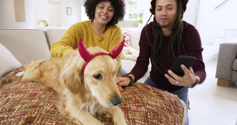 African american couple petting golden retriever wearing red horn headband in cozy home
