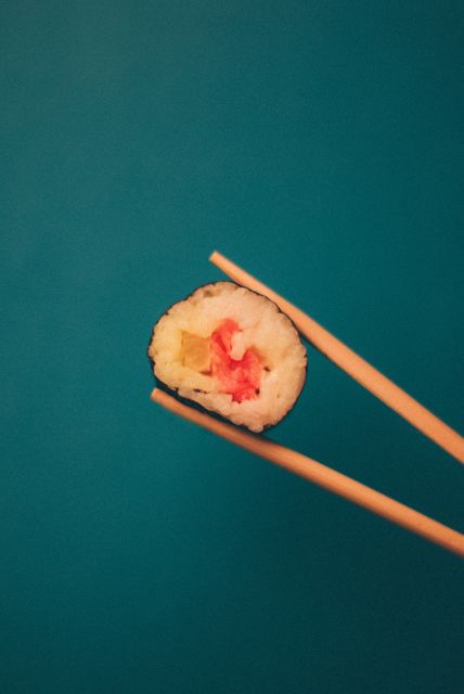 Sushi roll being held by a pair of chopsticks against a blue background. Ideal for use in food blogs, restaurant menus, culinary magazines, and promotional materials for Japanese cuisine. Highlighting Japanese culture and a healthy eating lifestyle.