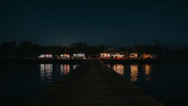 Wooden Pier Stretching Toward Lit Kiosks at Night with Warm Reflections on Calm Harbor