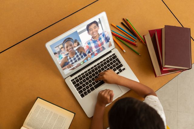 Child sitting at desk participating in a video call with friends using a laptop. Setup includes open books, colored pencils, and notebooks suggesting studying or homework environment. Suitable for illustrating remote learning, online education, children's social interaction, digital classroom concepts, e-learning platforms, and educational content.