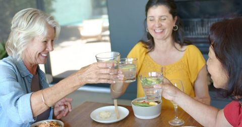 Diverse senior women clinking lemon water glasses during sunlit outdoor lunch gathering