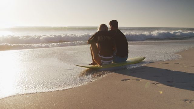 Couple enjoying romantic moments at sundown on beach, seated on surfboard while embracing each other. Gentle waves and evening sky create serene atmosphere depicting love and tranquility. Suitable for use in themes related to romance, vacations, beach lifestyle, travel advertising, and relationship bonding.