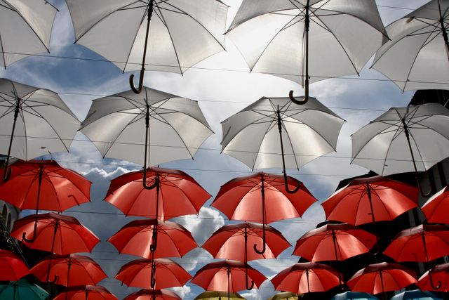 Numerous red and white umbrellas are suspended above an open pathway, creating a striking visual effect against a cloudy sky background. Useful for showcasing creative urban decorations, street festivals, and outdoor art installations. Ideal for use in travel blogs, cultural events promotions, and urban design showcases.