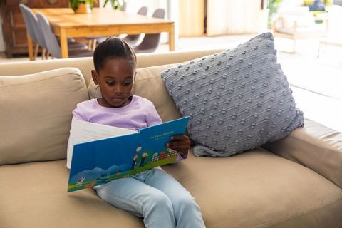 African american girl reading picture book on beige sofa in sunlit living room