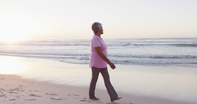 Senior woman walking on sandy beach with ocean waves in background during sunrise. Ideal for use in health and wellness campaigns, articles about active aging, promoting beach vacations, and depicting solitude and relaxation in nature.