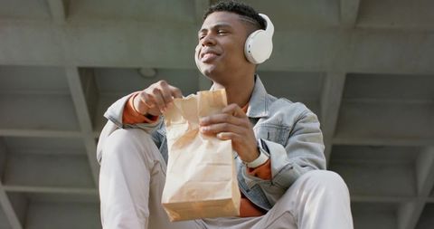 Young african american man sitting on concrete ledge enjoying lunch and listening to music