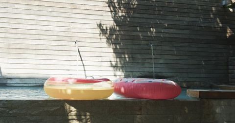 Sunlit Donut Floats Resting on Backyard Pool Coping beside Arcing Water Spouts