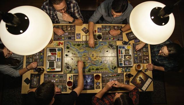 Six adults gathering around table playing strategic fantasy board game with colorful tokens