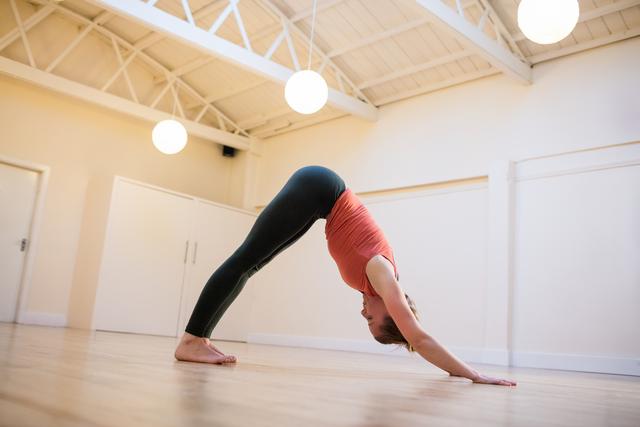 Woman practicing downward dog pose in a bright, spacious fitness studio with wooden floors and white walls. Ideal for use in articles or advertisements related to yoga, fitness, wellness, healthy lifestyle, and exercise routines. Can also be used in promotional materials for yoga studios, fitness centers, or wellness programs.