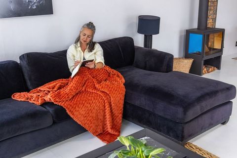 Mature woman journaling on navy chaise wrapped in rust blanket beside modern wood stove
