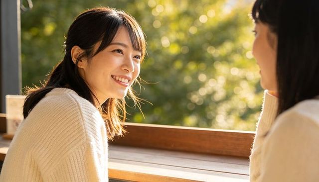 Sunlit asian woman smiling and chatting on terrace counter in cream knit sweater