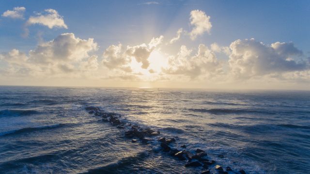 Sunlight beams through clouds over ocean with rock path in view. Ideal for travel websites, inspirational content, nature-themed projects, relaxation visuals, wallpapers, and backgrounds focusing on tranquility and natural beauty.
