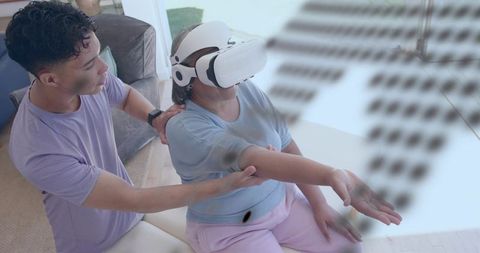 Young caregiver guiding senior using white vr headset during home rehab session