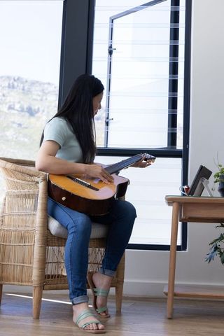 African american mature woman sitting in wicker armchair playing acoustic guitar by sunny window