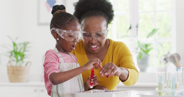 Grandmother and granddaughter wearing protective goggles conducting a science experiment together in a bright kitchen. The child is focused while the grandmother assists, creating a moment of bonding and learning. Suitable for use in educational contexts, family activities promotions, or highlighting intergenerational learning and STEM education.