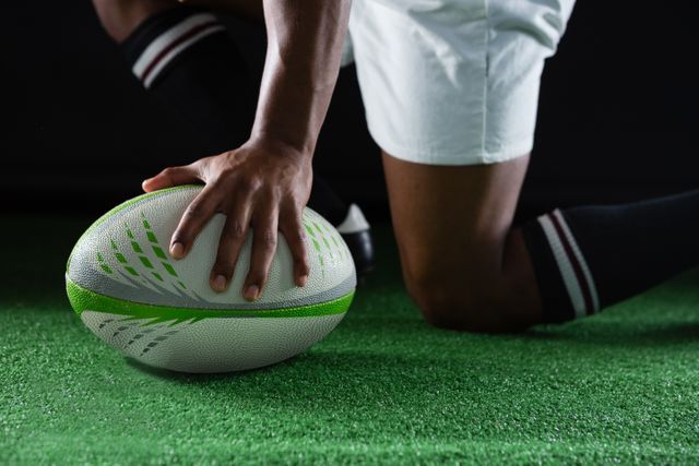 Rugby player kneeling on green grass field, holding rugby ball in preparation for game. Ideal for use in sports-related content, fitness promotions, team spirit campaigns, and athletic training materials.