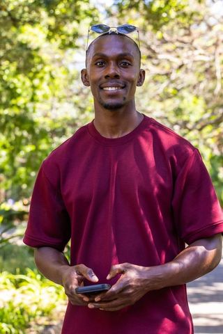 African american man smiling while holding smartphone outdoors in maroon shirt casual park