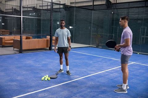 Two men playing padel on indoor blue court with lounge seating and glass walls