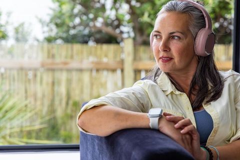 Midlife woman listening to music with pink headphones and smartwatch by window