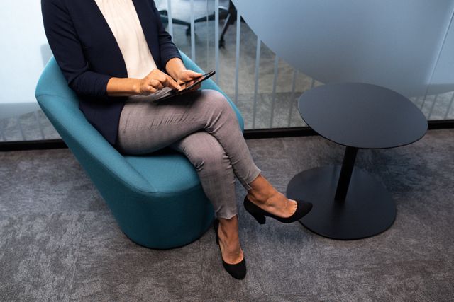 Businesswoman in formal attire sitting in a modern office lobby using a smartphone. Ideal for illustrating concepts related to business communication, professional environments, modern workplaces, and corporate settings. Useful for business websites, corporate blogs, and articles about office life or technology in business.