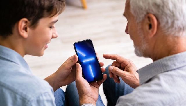 Grandfather teaching grandson smartphone use on couch, family technology bonding