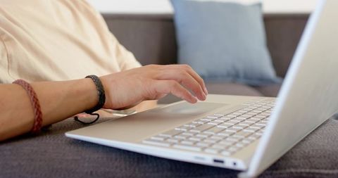 Asian man typing on silver laptop on sofa wearing braided wristbands for casual remote work