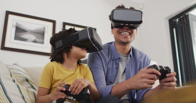 Father and son sitting on a sofa are experiencing virtual reality gaming together. Both wearing VR headsets, they appear to be fully engaged, holding controllers, smiling, and enjoying their time. This depiction of family bonding through technology can be used in ads, articles, or websites showcasing modern tech in home entertainment, family activities, and the impact of video games on relationships.