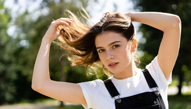 Sunlit young woman lifting hair in denim overalls and white tee, freckled natural portrait