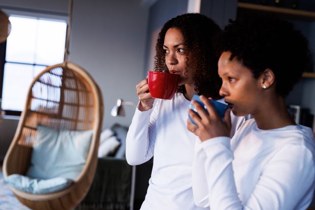 Happy, diverse lesbian couple drinking coffee and looking out of window standing in kitchen. Togetherness, free time and domestic life concept.