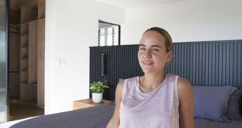 Smiling woman sitting on made bed in modern minimalist bedroom basking in natural light