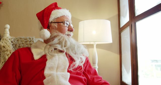 An elderly man dressed as Santa Claus is looking out of a window with a pensive expression, embodying the holiday spirit. Soft lighting from a nearby lamp creates a cozy, warm ambiance. This image is perfect for representing Christmas, holiday greetings, festive commercials, promotional materials, or nostalgic holiday moments.