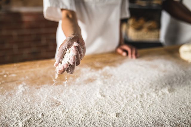 Midsection of Hispanic female baker holding flour over a wooden kitchen counter at a bakery. Ideal for use in articles or advertisements related to baking, culinary arts, food preparation, and the food and drink industry. Perfect for showcasing skilled labor, artisan baking, and the hands-on process of making baked goods.