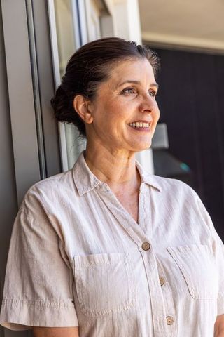 Serene mature woman standing by window smiling in beige button-up shirt lifestyle portrait