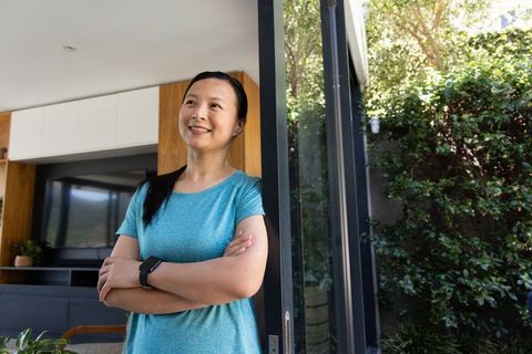 Mature asian woman standing at glass door enjoying garden view and morning sunlight
