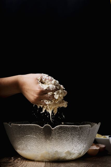 Hand preparing dough in large rustic bowl against dark background. Arm actively engaged in mixing ingredients, resulting in crumbs falling into bowl. Ideal for use in culinary blogs, cooking tutorials, food preparation articles, and advertisements for baking products or homemade cooking.