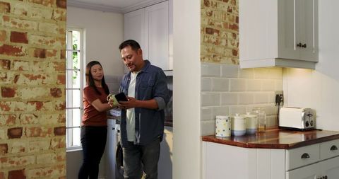 Asian couple checking smartphone while doing laundry in cozy kitchen with brick wall