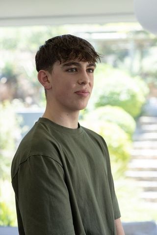 Teen male standing by window in olive t-shirt looking contemplative with garden background