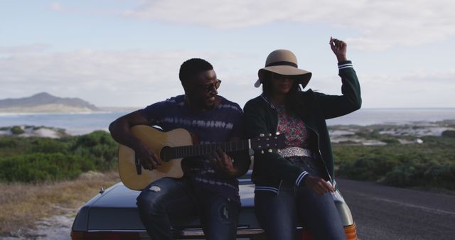 Diverse couple sitting on car by roadside playing guitar and singing. summer road trip on a country highway by the coast.