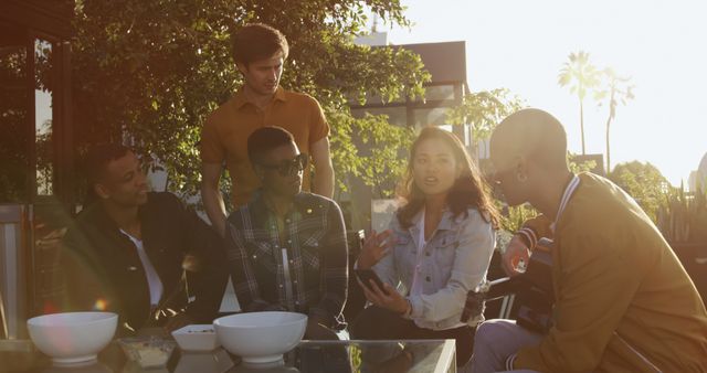 Diverse group of five friends gathered outdoors at sunset, engaging in lively conversation and laughter. Ideal for illustrating friendship, social interactions, cultural diversity, and outdoor leisure activities.