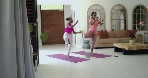 African mother and daughter practicing yoga tree pose on purple mats in modern living room