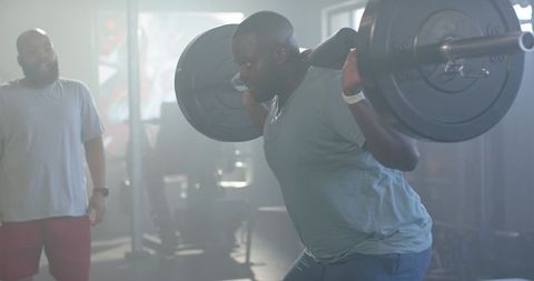 African man performing heavy barbell back squat while trainer spotting in gym workout