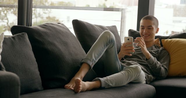 A young person relaxing on a comfortable couch while engaging with a smartphone. Ideal for promoting leisure activities, modern lifestyle, social media engagement, home comfort, and technology use in daily life.