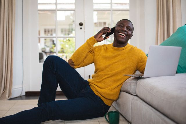 Young African American man sitting on floor, leaning against sofa, talking on smartphone while using laptop. Ideal for illustrating concepts of remote work, modern communication, home lifestyle, and technology use in everyday life.